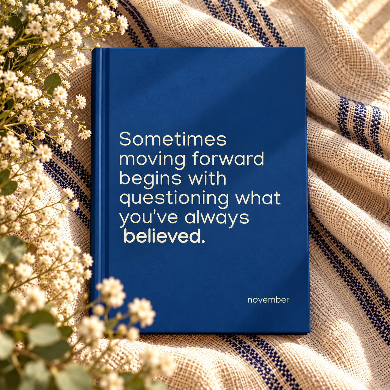 Blue hardcover "November" journal with cream motivational quote on striped blanket surrounded by white baby’s breath flowers