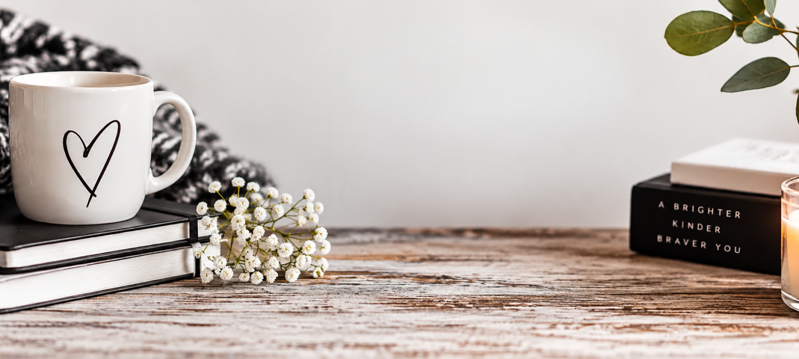 Warm inviting still life with heart coffee mug, stacked notebooks, baby’s breath blooms, candle and books on rustic table